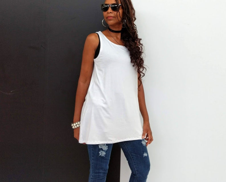 May include: A white sleeveless tunic top paired with distressed blue jeans. The top has a relaxed fit and a slightly asymmetrical hemline. The jeans feature ripped details at the knees.