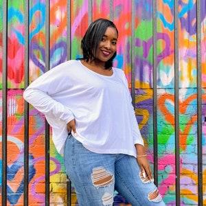 May include: A woman wearing a white long-sleeve shirt and ripped blue jeans. The background is a colorful graffiti wall with a variety of colors including pink, orange, green, and purple. The woman is smiling.