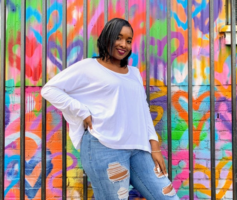 May include: A woman wearing a white long-sleeve shirt and ripped blue jeans. The background is a colorful graffiti wall with a variety of colors including pink, orange, green, and purple. The woman is smiling.