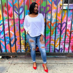 May include: A woman wearing a white long-sleeve shirt, distressed blue jeans, and red flats stands in front of a colorful graffiti wall. The jeans have multiple rips and tears. The wall features a vibrant mix of colors and patterns.