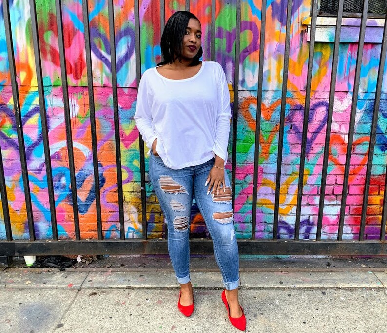 May include: A woman wearing a white long-sleeve shirt, distressed blue jeans, and red flats stands in front of a colorful graffiti wall. The jeans have multiple rips and tears. The wall features a vibrant mix of colors and patterns.