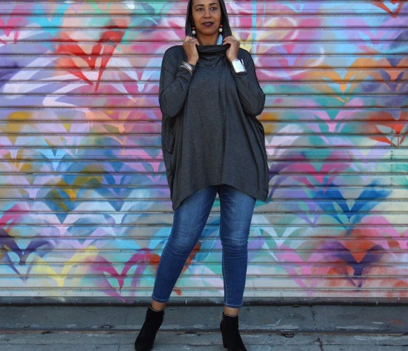 May include: A woman wearing a dark gray cowl-neck sweater, blue jeans, and black ankle boots. The background is a colorful mural with various shades of pink, blue, yellow, and purple. The sweater has long sleeves and a loose fit.