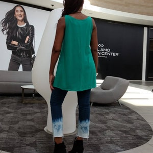 May include: A teal sleeveless tunic top paired with dark wash jeans featuring a white, faded, tie-dye effect at the ankles. The person is wearing black ankle boots. The background includes a large white sculpture and a shopping center.