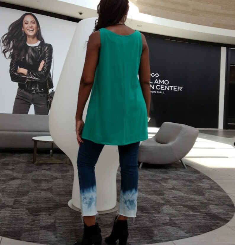 May include: A teal sleeveless tunic top paired with dark wash jeans featuring a white, faded, tie-dye effect at the ankles. The person is wearing black ankle boots. The background includes a large white sculpture and a shopping center.