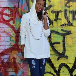 May include: A white long-sleeve top with a draped cowl neck and asymmetrical hemline. The top is paired with a long, oval-linked necklace and distressed blue jeans. The background is a colorful graffiti wall.