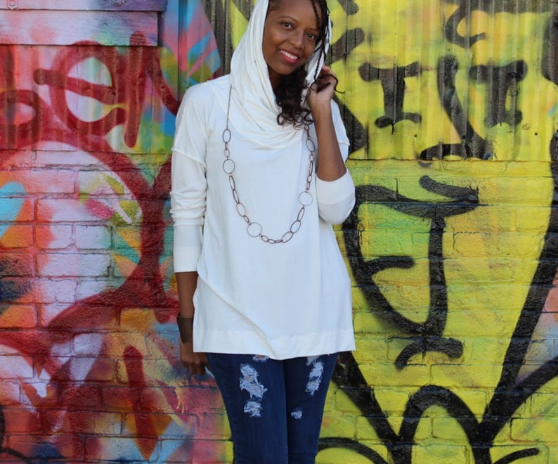 May include: A white long-sleeve top with a draped cowl neck and asymmetrical hemline. The top is paired with a long, oval-linked necklace and distressed blue jeans. The background is a colorful graffiti wall.