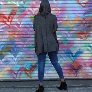 May include: A person wearing a dark gray hooded top with pockets and blue jeans. The person is standing in front of a colorful graffiti wall. The outfit includes black ankle boots.