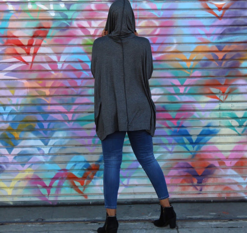 May include: A person wearing a dark gray hooded top with pockets and blue jeans. The person is standing in front of a colorful graffiti wall. The outfit includes black ankle boots.