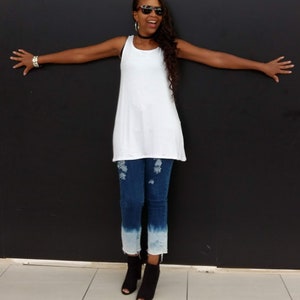 May include: White tank top with a scoop neck and a flowing silhouette. The top is paired with blue jeans featuring distressed details and a faded, ombre effect at the ankles. Black open-toe booties complete the outfit.