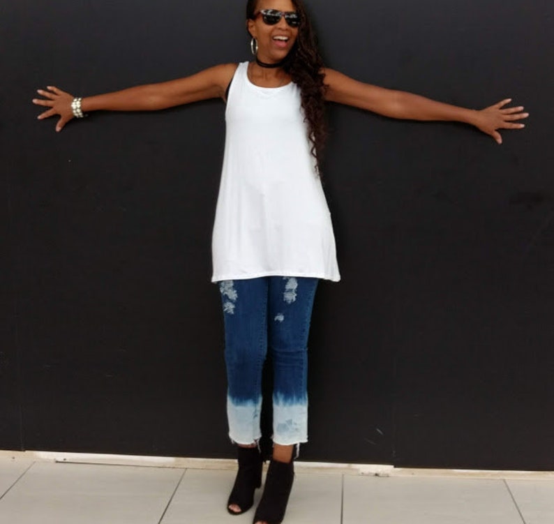 May include: White tank top with a scoop neck and a flowing silhouette. The top is paired with blue jeans featuring distressed details and a faded, ombre effect at the ankles. Black open-toe booties complete the outfit.