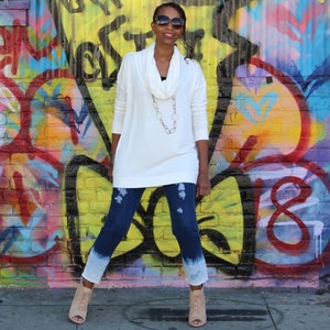 May include: A woman wearing a white cowl-neck sweater, distressed blue jeans with a white dip-dye effect, and beige open-toe booties. She is wearing sunglasses and a necklace. The background is a colorful graffiti wall.
