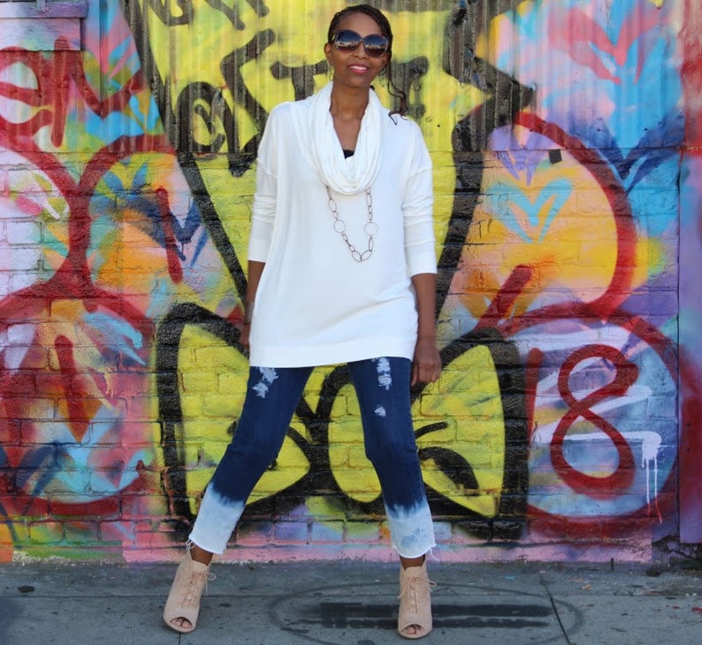 May include: A woman wearing a white cowl-neck sweater, distressed blue jeans with a white dip-dye effect, and beige open-toe booties. She is wearing sunglasses and a necklace. The background is a colorful graffiti wall.
