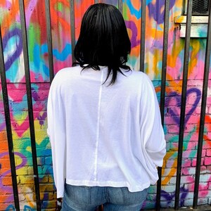 May include: A white long-sleeve shirt with a relaxed fit. The shirt is paired with blue jeans. The background features a colorful graffiti wall with a black metal fence.