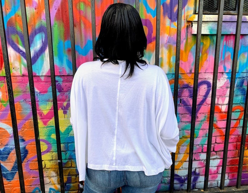 May include: A white long-sleeve shirt with a relaxed fit. The shirt is paired with blue jeans. The background features a colorful graffiti wall with a black metal fence.