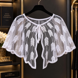 May include: A white sheer mesh capelet featuring a scalloped hem and a repeating pattern of white sequined leaf motifs. The garment has a round neckline with a delicate tie closure at the center front, constructed from lightweight, translucent fabric.