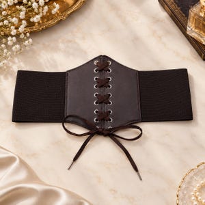 May include: A wide black elastic corset belt featuring a central dark brown faux leather panel with silver-toned metal eyelets and a crisscross lace-up design. The front includes a decorative brown ribbon tied into a bow at the bottom.