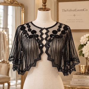 Puede incluir: Una capa bolero de malla negra transparente con intrincados abalorios negros y patrones florales de lentejuelas. La prenda presenta un dobladillo festoneado, un cuello redondo y un diseño central de corte en zigzag con flecos de cuentas.