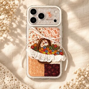 May include: A clear smartphone case featuring a patchwork floral background in shades of peach, orange, and burgundy. An embroidered basket filled with colorful flowers and a white lace trim is attached, accented by a thin white ribbon tied in a bow.