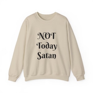May include: A beige crewneck sweatshirt featuring the black text "NOT Today Satan" printed in a stylized serif font across the center of the chest. The garment has long sleeves with ribbed cuffs and a ribbed neckline, providing a simple design.