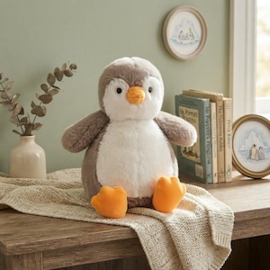 May include: A plush penguin toy with soft grey and white fur, featuring a bright orange beak and orange feet. The stuffed animal is seated upright, showcasing a rounded body, small black eyes, and a textured, fuzzy appearance throughout its design.