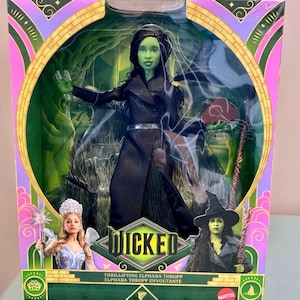 May include: A Mattel Wicked doll of Elphaba Thropp in a black coat with a silver belt, packaged in a box featuring the Wicked logo, an image of Glinda with a wand, and an image of Elphaba with a broomstick against a green forest background.