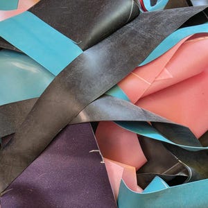 May include: A collection of overlapping, flexible strips of material in black, light blue, and soft pink. The strips are smooth and appear to be made of a pliable, non-porous substance, layered in a disorganized pile with varying widths and lengths.