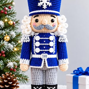 Pode incluir: Um boneco quebra-nozes de crochê com uniforme azul e detalhes brancos, seis botões brancos e um floco de neve branco no chapéu. O boneco tem cabelo branco encaracolado, bigode cinza, calças cinzas e botas pretas com cadarços brancos.
