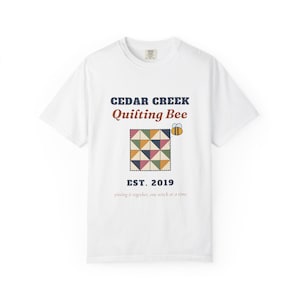 May include: A white t-shirt featuring the text CEDAR CREEK Quilting Bee EST. 2019 and piecing it together, one stitch at a time. The graphic includes a square quilt made of multi-colored triangles and a small yellow and black bee.