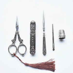 May include: A silver sewing set featuring ornate scissors with a brown tassel, a cylindrical needle case with floral embossing, a pointed stiletto tool, a thin flat needle threader, and a dimpled thimble, all arranged on a plain white surface.