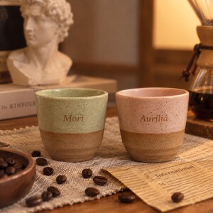Puede incluir: Dos tazas de cerámica con acabado moteado, una en verde claro y otra en rosa claro. Cada taza presenta una base marrón natural sin esmaltar con ranuras horizontales y los nombres Mori y Aurilia grabados en el centro de las secciones.