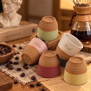 Puede incluir: Un conjunto de cinco tazas de cerámica con diseño bicolor, que presenta una base marrón natural y una sección superior coloreada en verde claro, rosa, blanco, rojo oscuro y amarillo. Dos tazas están personalizadas con los nombres Selene y Kael.