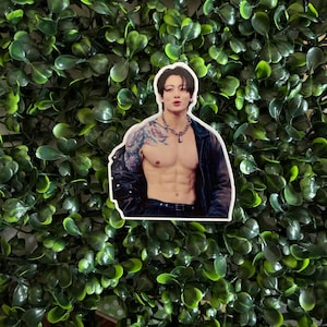 May include: A die-cut sticker featuring a person with dark hair, a silver chain necklace, and a visible arm tattoo. The person is wearing an open dark jacket, revealing a bare chest and torso. The sticker has a white border.