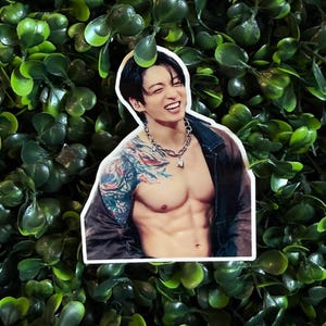 May include: A die-cut sticker featuring a smiling person with dark hair, wearing a dark open jacket and a silver chain necklace. The person has a large, colorful tattoo covering their right shoulder and upper arm, and a visible muscular torso.