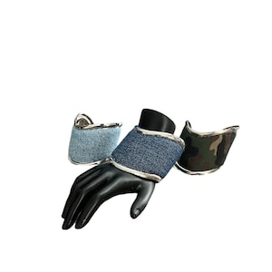 Può includere: Tre braccialetti in tessuto con bordatura in metallo argentato. Uno è in denim blu chiaro, uno in denim blu scuro e uno in mimetico.