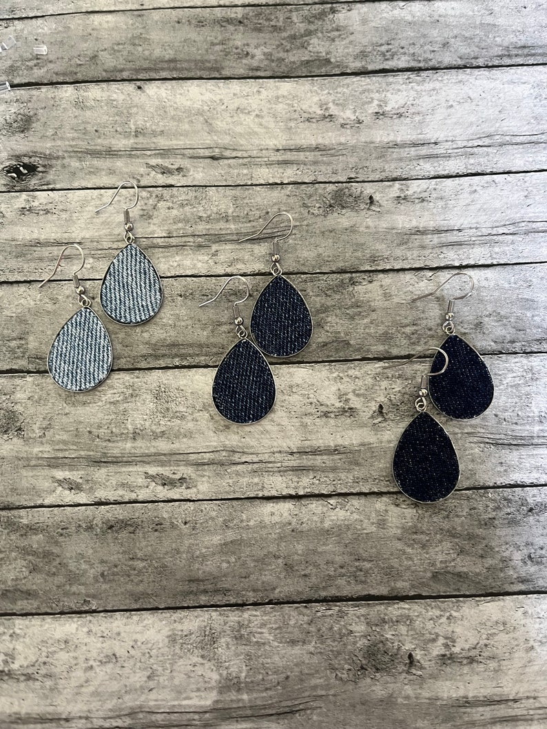 May include: Three pairs of denim teardrop earrings. The earrings are silver with a denim fabric covering the teardrop shape. One pair is light blue denim, one pair is dark blue denim, and one pair is black denim.