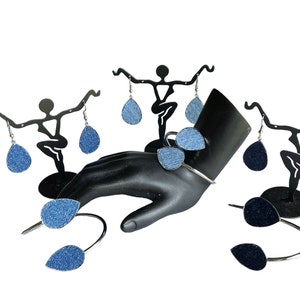 May include: A set of denim teardrop earrings displayed on a black mannequin hand and two black earring stands. The earrings are a light blue denim fabric.
