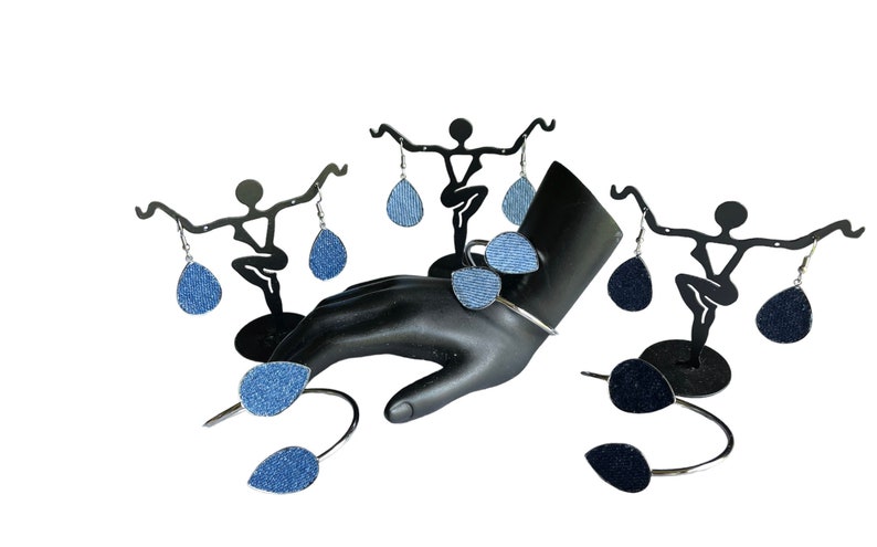 May include: A set of denim teardrop earrings displayed on a black mannequin hand and two black earring stands. The earrings are a light blue denim fabric.