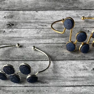 May include: Two silver and gold bracelets with blue fabric covered buttons. The bracelets are on a wooden surface.
