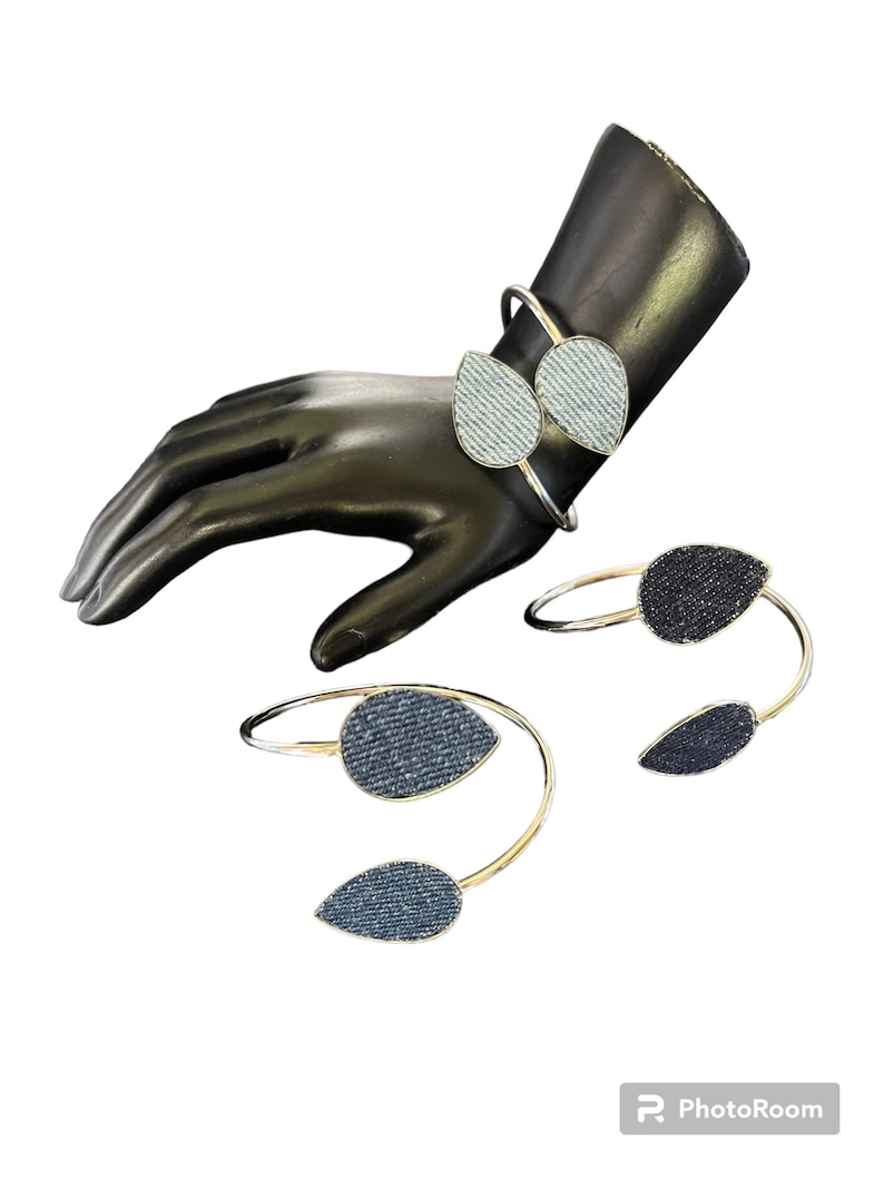 May include: Three gold bangle bracelets with denim fabric accents. The bracelets are in the shape of a teardrop and are stacked on a mannequin's wrist.