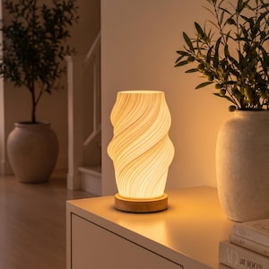 Puede incluir: Una lámpara de mesa cilíndrica con una pantalla translúcida de color blanco roto con textura en espiral que emite un brillo cálido. La lámpara cuenta con una base circular de madera clara y se asienta sobre una superficie plana.
