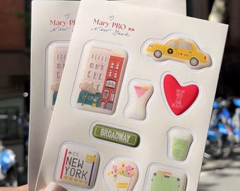 May include: A sticker sheet labeled Mary PRO New York featuring various designs including a yellow taxi, a red building, a martini glass, a red heart with text, a green Broadway sign, a coffee cup, a New York sign, a flower bouquet, and a suitcase.