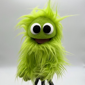 May include: A bright lime green hand puppet featuring long, shaggy faux fur, a rounded felt face, and two large white eyes with black pupils. The puppet has a wide, open mouth with a dark interior, creating a distinct character appearance.