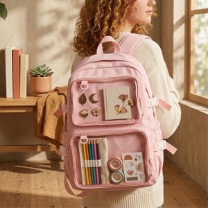Puede incluir: Una mochila de color rosa claro con dos bolsillos frontales transparentes. El bolsillo superior muestra cuatro pines pequeños y un cuaderno con arte floral. El bolsillo inferior contiene lápices de colores, dos rollos de cinta y pegatinas.