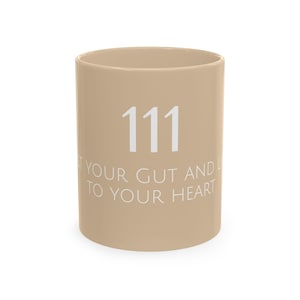 May include: A light tan ceramic mug featuring the number 111 in large white font at the top, followed by the white text "TRUST YOUR GUT AND LISTEN TO YOUR HEART" printed in a clean, sans-serif typeface across the center of the cylindrical body.