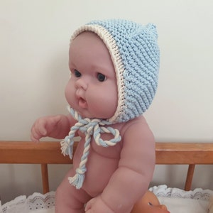 May include: A light blue and cream knitted baby bonnet with a tie fastening. The bonnet is made from soft yarn and has a ribbed texture. The tie is a twisted cord in matching colours. The bonnet is designed to fit a baby.