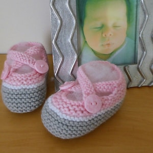 May include: A pair of knitted baby shoes in pink and grey with white trim. The shoes have a strap and a button fastening.
