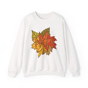 May include: A white long-sleeve crewneck sweatshirt featuring a central graphic of colorful autumn leaves in shades of red, orange, yellow, and green. The word FALL is printed in a simple, dark brown sans-serif font over the leaf cluster.