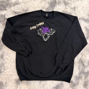 May include: A black long-sleeve sweatshirt featuring a graphic of three white ghosts and the purple text "Basic Witch" centered on the chest. Above the ghosts, a gold-colored script logo reads "Kroger Creations" in a cursive font style.