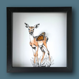 May include: A black framed illustration of a fawn with a monarch butterfly on its back. The fawn is standing in a field of grass.