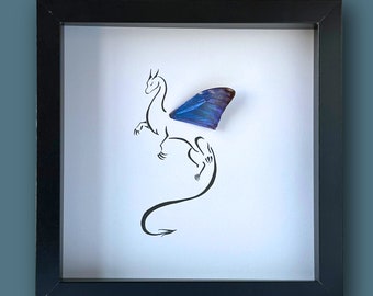 Pode incluir: Uma caixa de sombra enquadrada em preto com uma asa de borboleta azul montada em um fundo branco. A asa de borboleta está posicionada para parecer a asa de um dragão. O dragão está delineado em preto.